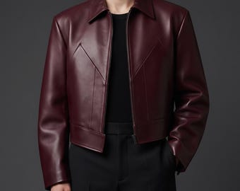 May include: A deep burgundy leather jacket featuring a classic collar, a full-length front zipper, and distinct V-shaped seam detailing across the chest. The jacket is worn over a plain black shirt and paired with black trousers, creating a sleek look.