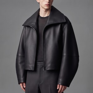 May include: A black leather jacket featuring a wide, oversized collar and a front zipper closure. The jacket has long, loose sleeves and a cropped hemline, worn over a dark charcoal crew neck shirt and matching dark charcoal trousers.