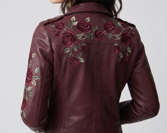 May include: A burgundy leather jacket featuring intricate floral embroidery on the back and sleeves. The design showcases deep red roses with green leaves, a classic collar, shoulder epaulets, and a fitted silhouette with visible seam detailing.