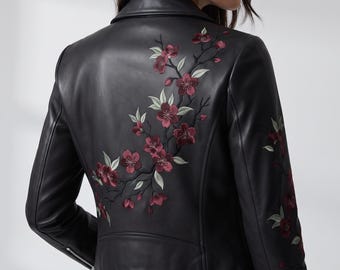 May include: A black leather jacket featuring an embroidered floral design of dark red cherry blossoms with light green leaves extending across the back and down the right sleeve, complete with a notched collar and silver-toned metal zipper details.
