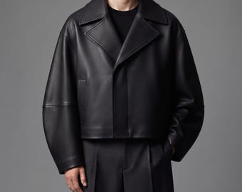 May include: A black cropped jacket with a smooth finish, featuring a wide notched collar, long sleeves with dropped shoulders, and a concealed front closure. The jacket is worn over a plain black shirt and paired with dark grey tailored trousers.