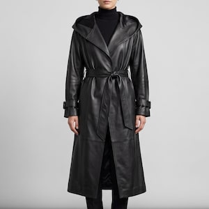 May include: A black leather trench coat featuring a large hood, long sleeves with buckled cuffs, and a matching tie belt at the waist. The coat has a structured collar, a front button closure, and a long, straight silhouette that reaches below the knees.