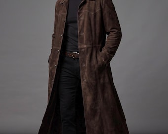 May include: A long, dark brown suede coat featuring a classic collar, a button-front closure, and vertical seam detailing. The coat is worn over a black t-shirt and black trousers, with a brown leather belt visible at the waistline.