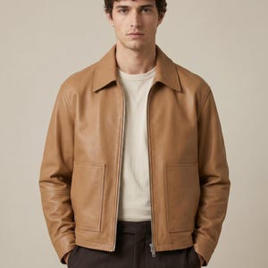 May include: A tan leather jacket with a collared neckline and a front zipper closure. The jacket has two front pockets and long sleeves. The model is wearing a white t-shirt underneath the jacket.