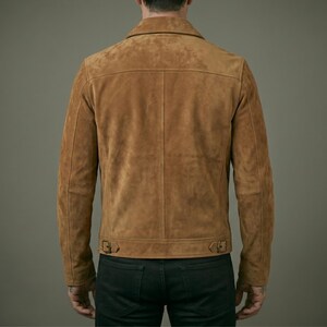 May include: A tan suede jacket featuring a pointed collar, long sleeves, and a horizontal back yoke. The hem includes two adjustable side tabs with metal buckles. The jacket is worn over dark pants, showcasing a structured, clean-lined design.