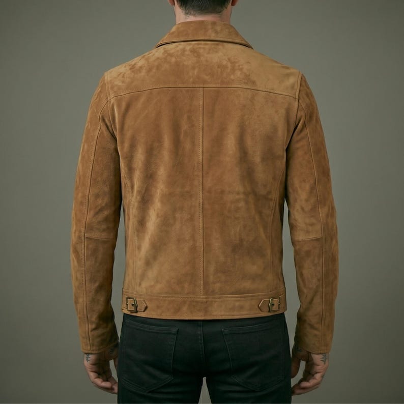 May include: A tan suede jacket featuring a pointed collar, long sleeves, and a horizontal back yoke. The hem includes two adjustable side tabs with metal buckles. The jacket is worn over dark pants, showcasing a structured, clean-lined design.