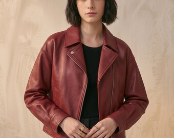 May include: A deep red leather jacket featuring a wide notched collar, long sleeves with cuffs, and a silver-toned metal zipper closure. The jacket is worn over a black ribbed top, and the design includes a single silver snap button at the collar.