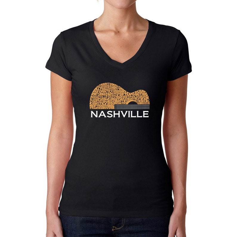 Puede incluir: Camiseta negra de cuello en V con un gr&aacute;fico de una silueta de guitarra compuesta por palabras relacionadas con Nashville, Tennessee. Las palabras son de color naranja y la guitarra est&aacute; delineada en gris. El texto "NASHVILLE" est&aacute; impreso en blanco debajo de la guitarra.