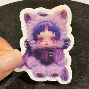 May include: A sticker featuring a doll-like figure in a purple furry outfit with a hood. The figure has blue hair, large eyes, and a small horn. The sticker has a white border and is held by a hand.
