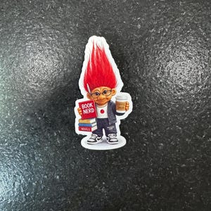 May include: A sticker featuring a cartoon troll with bright red hair, glasses, and a jacket. The troll holds books labeled "BOOK NERD" and "NOVELS" and a coffee cup that says "COFFEE SNOB".