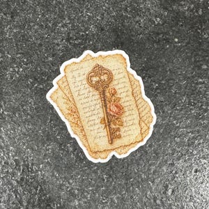 Op de afbeelding: Een sticker met een vintage sleutelontwerp, een roos en tekst op verouderd papier. De sleutel is goudkleurig en het papier heeft een sepia-tint. De sticker heeft een witte rand op een donkergrijze achtergrond.