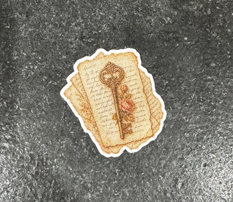 Op de afbeelding: Een sticker met een vintage sleutelontwerp, een roos en tekst op verouderd papier. De sleutel is goudkleurig en het papier heeft een sepia-tint. De sticker heeft een witte rand op een donkergrijze achtergrond.