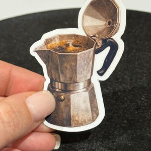 May include: A sticker of a vintage-style stovetop espresso maker, also known as a Moka pot. The sticker is a sepia tone with a black handle and a lid that is open. The coffee pot is filled with coffee.