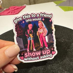 May include: A sticker featuring two doll figures with pink face coverings and holding shovels. The sticker has the text "give this to a friend who would show up without asking" in a pink and white font.