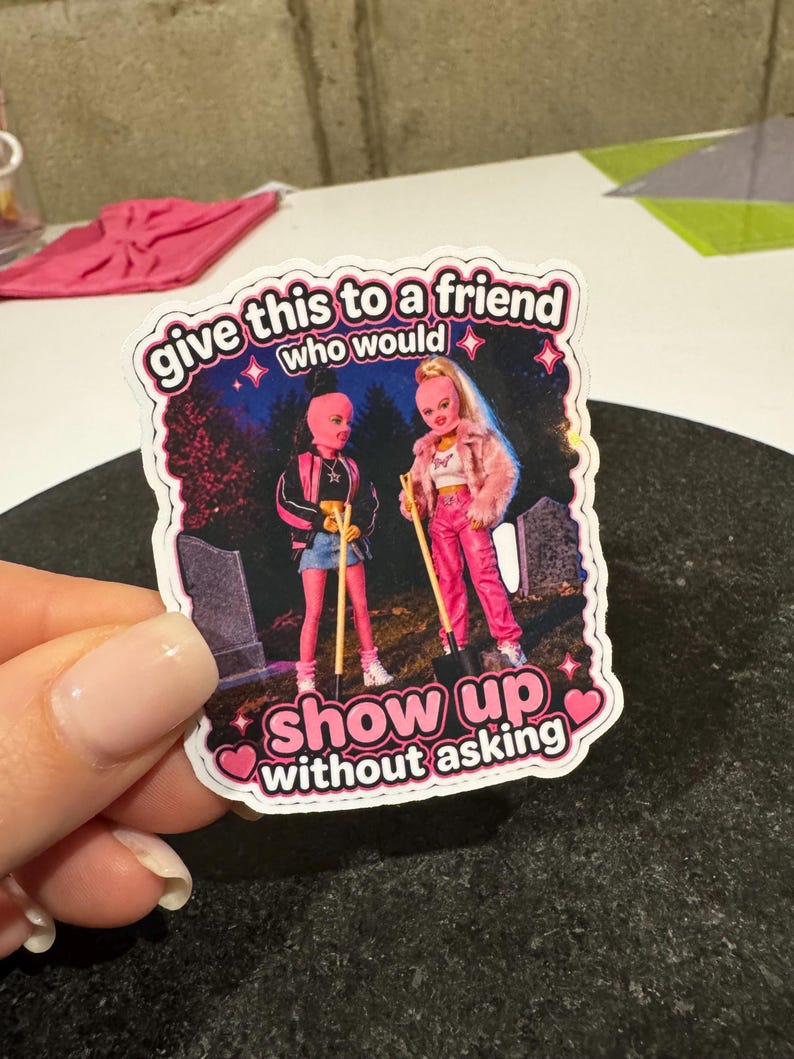 May include: A sticker featuring two doll figures with pink face coverings and holding shovels. The sticker has the text "give this to a friend who would show up without asking" in a pink and white font.