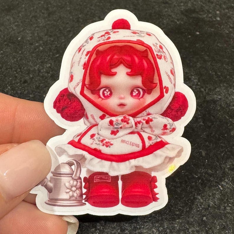K&ouml;nnte beinhalten: Ein Aufkleber mit einer puppenartigen Figur mit roten Haaren und passendem Outfit. Die Figur tr&auml;gt ein wei&szlig;es Kleid mit roten Blumenmustern, eine Kapuze und rote Stiefel. Eine kleine Gie&szlig;kanne ist ebenfalls im Design enthalten.