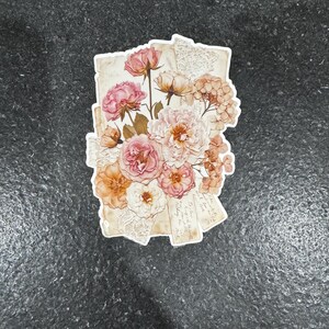 Op de afbeelding: Een sticker met een boeket van roze, perzik en witte bloemen. De bloemen zijn gerangschikt op papier in vintage-stijl met kanten details. De sticker heeft een witte rand en staat op een donkergrijze achtergrond.