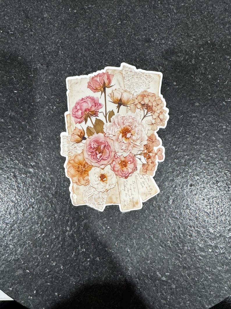 Op de afbeelding: Een sticker met een boeket van roze, perzik en witte bloemen. De bloemen zijn gerangschikt op papier in vintage-stijl met kanten details. De sticker heeft een witte rand en staat op een donkergrijze achtergrond.
