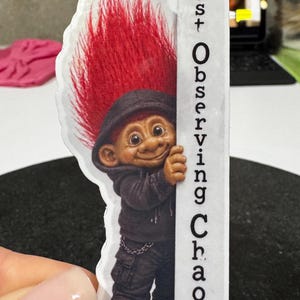 May include: A sticker featuring a troll with red hair and a black hoodie, smiling and peeking from behind a white sign. The sign reads "Just Observing Chaos."
