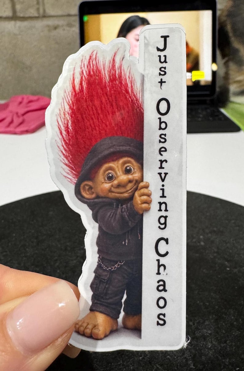 May include: A sticker featuring a troll with red hair and a black hoodie, smiling and peeking from behind a white sign. The sign reads "Just Observing Chaos."