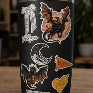 May include: Black tumbler covered in fantasy-themed stickers. Stickers include a dragon rider, a dragon with a smaller dragon, a mountain scene, a banner that says "BASGIATH FLY OR DIE", a heart that says "XADDY", and a bow that says "never again Valkyrie".