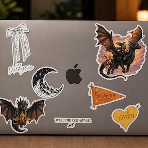 May include: A collection of fantasy-themed stickers on a laptop. Designs include a dragon with a rider, a crescent moon over mountains, a bow with text, a heart with an arrow, and a pennant flag. The text includes "Valkyrie", "BASGIATH", "FLY OR DIE", "XADDY", and "WELL THE F*CK AWARE."