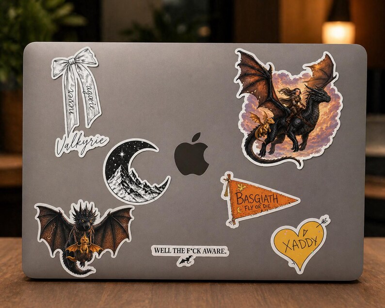 May include: A collection of fantasy-themed stickers on a laptop. Designs include a dragon with a rider, a crescent moon over mountains, a bow with text, a heart with an arrow, and a pennant flag. The text includes "Valkyrie", "BASGIATH", "FLY OR DIE", "XADDY", and "WELL THE F*CK AWARE."