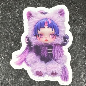 K&ouml;nnte beinhalten: Ein Aufkleber mit einer stilisierten Illustration einer puppenartigen Figur in einem lila Fellkost&uuml;m. Die Figur hat blaue Haare, gro&szlig;e Augen und einen kleinen Mund. Das Kost&uuml;m hat eine Kapuze und einen Knopfverschluss.