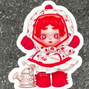 K&ouml;nnte beinhalten: Ein Aufkleber mit einer puppenartigen Figur mit roten Haaren und passendem Outfit. Die Figur tr&auml;gt ein wei&szlig;es Kleid mit roten Blumenmustern und einer passenden Kapuze. Eine kleine Gie&szlig;kanne ist ebenfalls im Design enthalten.