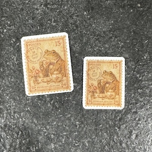 Op de afbeelding: Twee vintage postzegels. Elke postzegel toont een kikker in een pak die thee drinkt, met de tekst "Postes" en "R&eacute;publique Fran&ccedil;aise". De postzegels zijn bruin met witte randen.