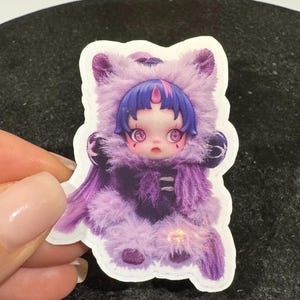 K&ouml;nnte beinhalten: Ein Aufkleber mit einer puppenartigen Figur in einem lila Fellkost&uuml;m mit Kapuze. Die Figur hat blaue Haare, gro&szlig;e Augen und ein rosa Mal auf der Stirn. Der Aufkleber wird von einer Hand vor einem dunklen Hintergrund gehalten.