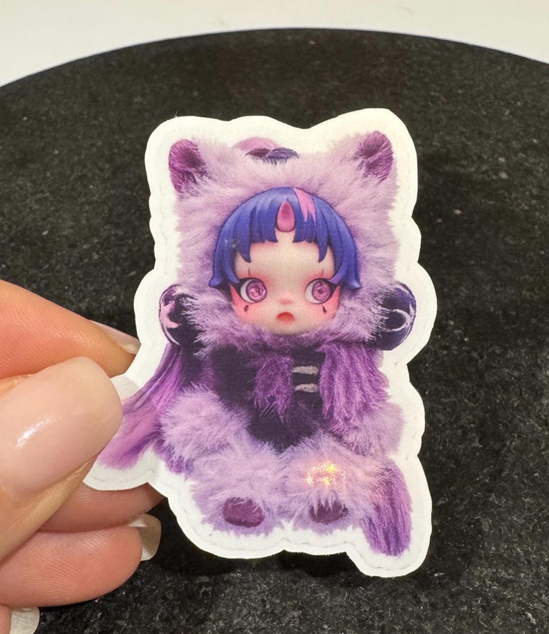 K&ouml;nnte beinhalten: Ein Aufkleber mit einer puppenartigen Figur in einem lila Fellkost&uuml;m mit Kapuze. Die Figur hat blaue Haare, gro&szlig;e Augen und ein rosa Mal auf der Stirn. Der Aufkleber wird von einer Hand vor einem dunklen Hintergrund gehalten.