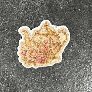 Op de afbeelding: Een sticker met een vintage theepotontwerp. De beige theepot is versierd met een bloemen- en vlindermotief en roze rozen. De sticker heeft een witte rand en is ideaal voor het decoreren van diverse items.