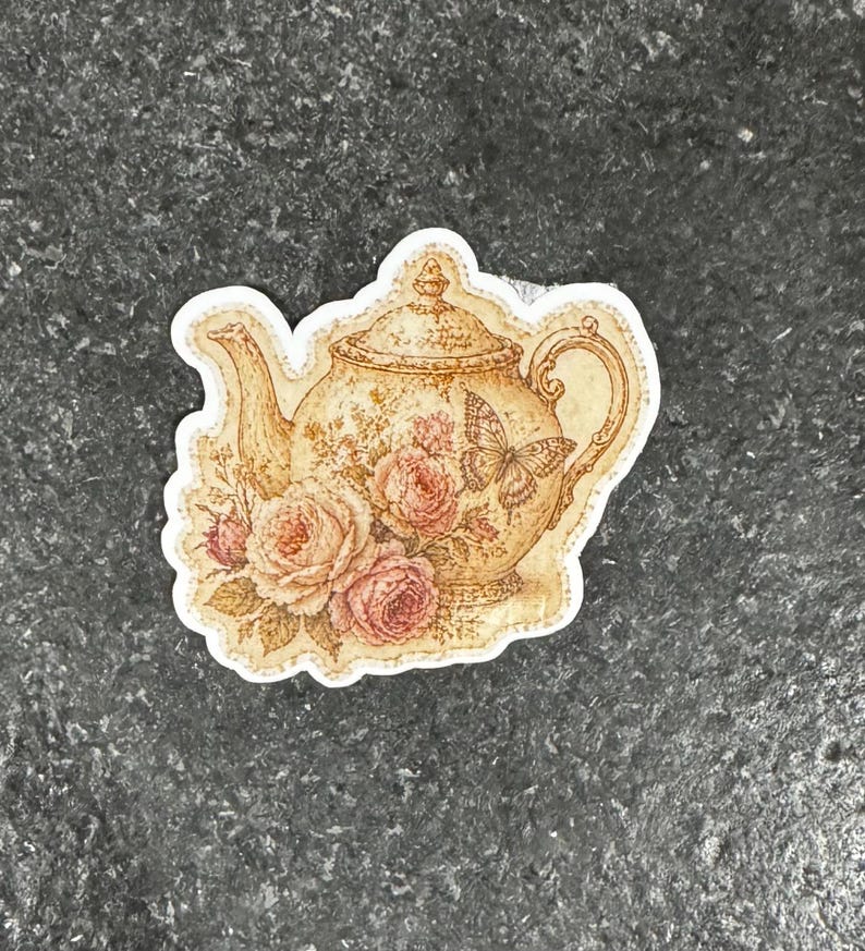 Op de afbeelding: Een sticker met een vintage theepotontwerp. De beige theepot is versierd met een bloemen- en vlindermotief en roze rozen. De sticker heeft een witte rand en is ideaal voor het decoreren van diverse items.