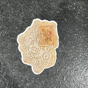 Op de afbeelding: Een vintage sticker met een cr&egrave;mekleurig kantpatroon en een postzegelontwerp. De postzegel toont een roos en de tekst "Postes" en "Paris 18 Juin 1907". De sticker heeft een witte rand.