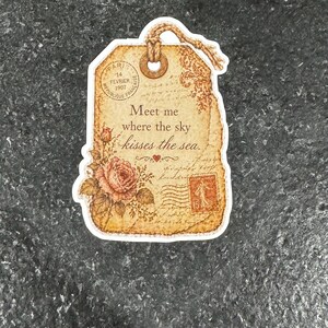 May include: A vintage-style tag with a distressed paper appearance. The tag features a rose illustration, a postage stamp, and the text "Meet me where the sky kisses the sea." It includes the text "PARIS 14 FEVRIER 1907".
