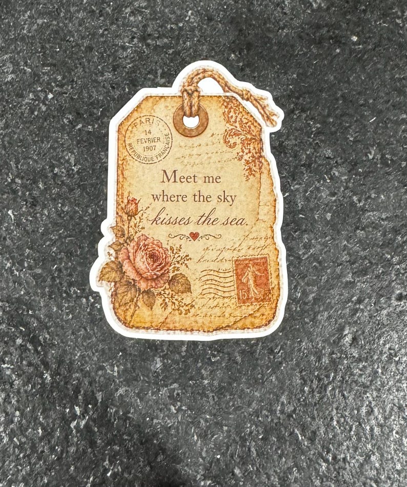 May include: A vintage-style tag with a distressed paper appearance. The tag features a rose illustration, a postage stamp, and the text "Meet me where the sky kisses the sea." It includes the text "PARIS 14 FEVRIER 1907".