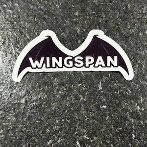 K&ouml;nnte beinhalten: Ein Aufkleber mit einem Design aus dunkelvioletten Fl&uuml;geln und wei&szlig;en Umrissen. Das Wort "WINGSPAN" steht in fetten wei&szlig;en Buchstaben in der Mitte. Der Aufkleber befindet sich auf einer strukturierten, dunkelgrauen Oberfl&auml;che.