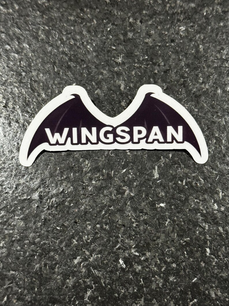 K&ouml;nnte beinhalten: Ein Aufkleber mit einem Design aus dunkelvioletten Fl&uuml;geln und wei&szlig;en Umrissen. Das Wort "WINGSPAN" steht in fetten wei&szlig;en Buchstaben in der Mitte. Der Aufkleber befindet sich auf einer strukturierten, dunkelgrauen Oberfl&auml;che.
