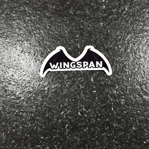 K&ouml;nnte beinhalten: Ein wei&szlig;er Aufkleber mit einem schwarzen Fledermausfl&uuml;gel-Design und dem Wort "WINGSPAN" in wei&szlig;er, fetter Schrift. Der Aufkleber befindet sich auf einer dunklen, strukturierten Oberfl&auml;che.