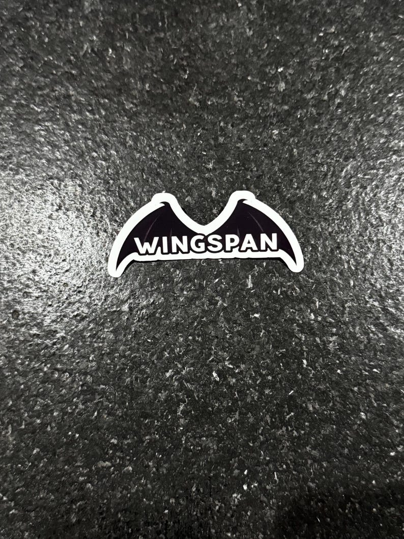K&ouml;nnte beinhalten: Ein wei&szlig;er Aufkleber mit einem schwarzen Fledermausfl&uuml;gel-Design und dem Wort "WINGSPAN" in wei&szlig;er, fetter Schrift. Der Aufkleber befindet sich auf einer dunklen, strukturierten Oberfl&auml;che.