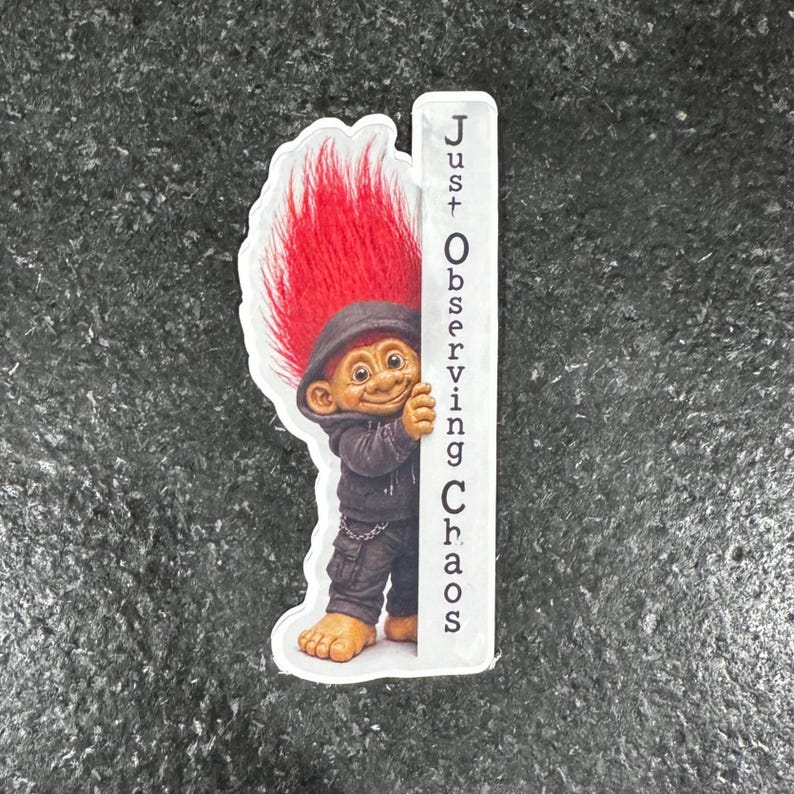May include: A sticker featuring a troll with red hair and a black hoodie, holding a white sign with the text "Just Observing Chaos" in black. The troll has a smiling expression and is standing on a gray surface.