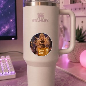 K&ouml;nnte beinhalten: Ein wei&szlig;er Stanley-Becher mit Griff und transparentem Deckel. Ein runder Aufkleber zeigt einen Anime-Charakter mit Fl&uuml;geln, den Text "PLAYER ONE" und ein Fragezeichen.