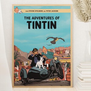 May include: A framed poster featuring The Adventures of Tintin with text from Steven Spielberg and Peter Jackson. The illustration shows Tintin, Captain Haddock, and Snowy in a sidecar motorcycle driving through a desert town with a bird flying above.