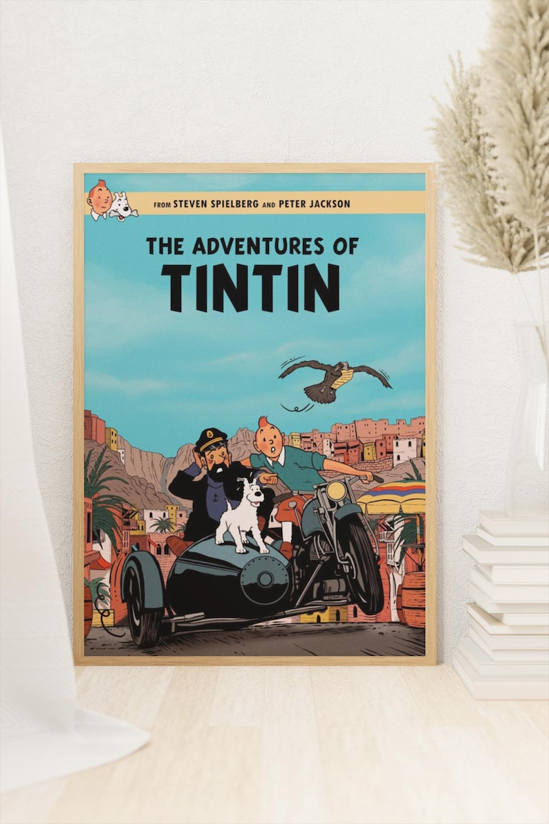 May include: A framed poster featuring The Adventures of Tintin with text from Steven Spielberg and Peter Jackson. The illustration shows Tintin, Captain Haddock, and Snowy in a sidecar motorcycle driving through a desert town with a bird flying above.