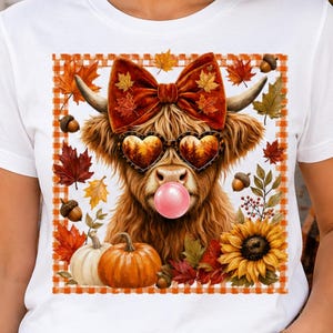 May include: A white short-sleeve t-shirt featuring a graphic of a Highland cow wearing heart-shaped sunglasses and a bow, blowing a pink bubble. The design includes autumn leaves, pumpkins, and a sunflower, all framed by an orange gingham border.