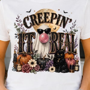 May include: A white short-sleeve t-shirt featuring a graphic print of a ghost wearing sunglasses and blowing a pink bubble, a black cat, pumpkins, flowers, and candles. The text reads "CREEPIN' IT REAL" in a stylized black font with a moon background.