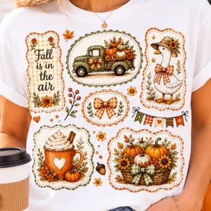 Op de afbeelding: Een wit t-shirt met een grafische print van een groene vrachtwagen met pompoenen, een eend met een hoed, een mok warme chocolademelk en een mand met pompoenen. De tekst "Fall is in the air" staat in een decoratief kader op de linkerzijde.