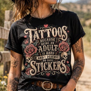 May include: Black t-shirt with the text "Tattoos because being an adult is hard and I deserve to have stickers." The shirt features a floral and skull design in pink and cream colors. A brown belt with a gold buckle is also visible.