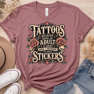 May include: Dusty rose t-shirt with a graphic that reads "Tattoos because being an adult is hard and I deserve to have stickers." The design includes roses, skulls, and hearts. The shirt is made of a soft material.